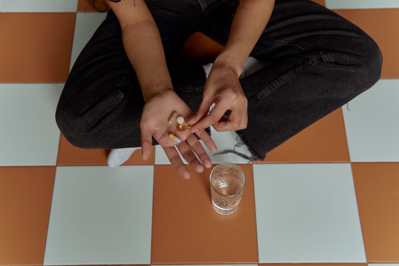 Individual sitting on checkered floor holding pills with a glass of water nearby.